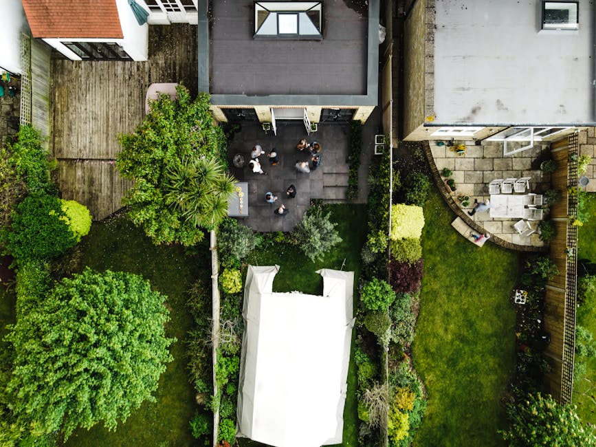 An aerial view of a small backyard garden showing a well-maintained lawn bordered by flower beds and shrubs. In the center, there is a paved patio area with several people gathered, possibly engaging 